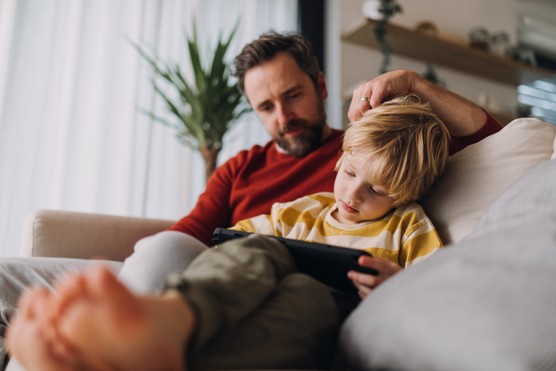Father and son sitting on a sofa together, looking at a tablet the boy holds.