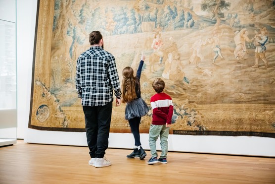 A young girl exploring a museum with her family and excitedly pointing up towards a large painting on display.
