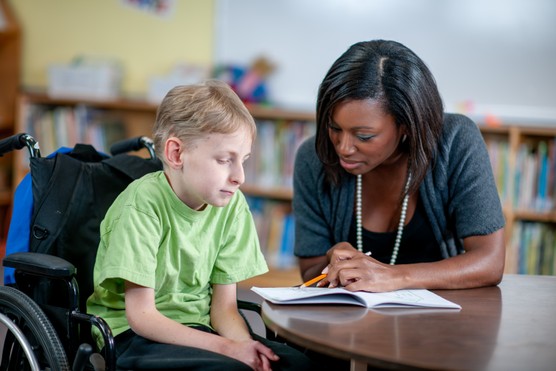 A young boy in a wheelchair works with an Educational Assistant on his school work. The boy is listening attentively as the worker helps him understand his studies.
